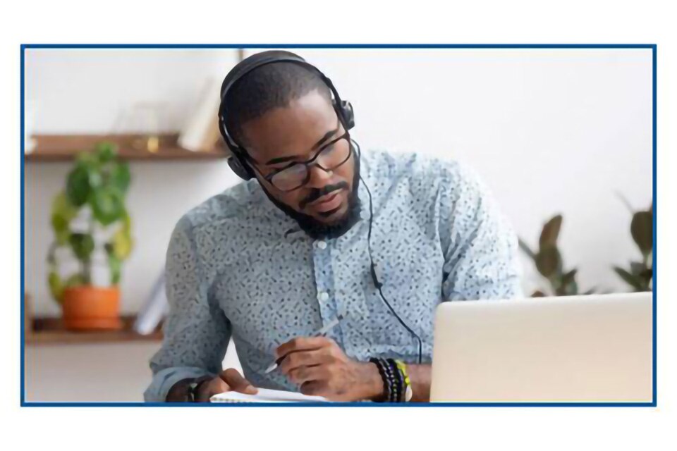 A man wearing headphones plugged into a laptop taking down notes