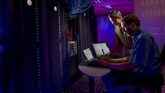 Two men working on laptops inside a server room