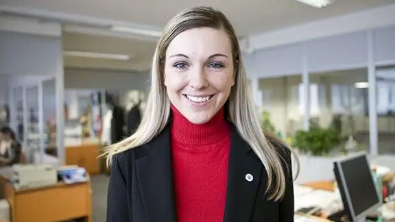 A woman standing in an office, looking into the camera and smiling