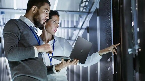 A man holding a laptop. standing next to a woman pointing at a server unit